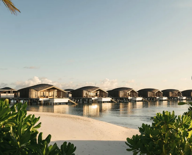Vilas sobre a água do Club Med Finolhu ao pôr do sol, com tetos curvos iluminados e areia branca